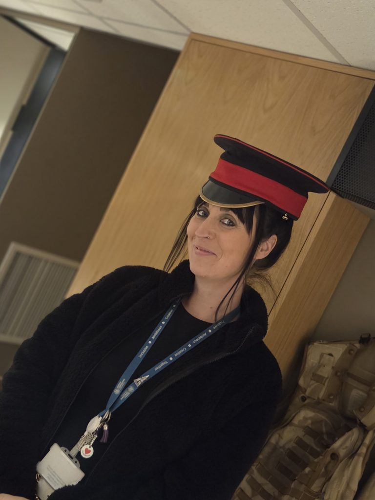A person sits indoors wearing a black graduation-style gown, lanyard and a red and black academic cap, smiling slightly towards the camera.