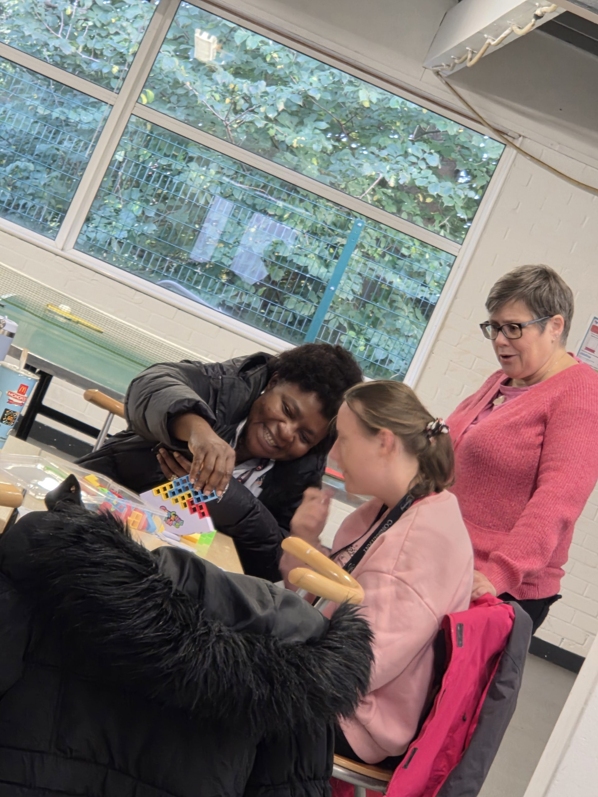 Three people take part in a creative group activity at a table in a bright community room. One person is helping another with colourful craft materials, while a third person stands nearby watching and supporting.