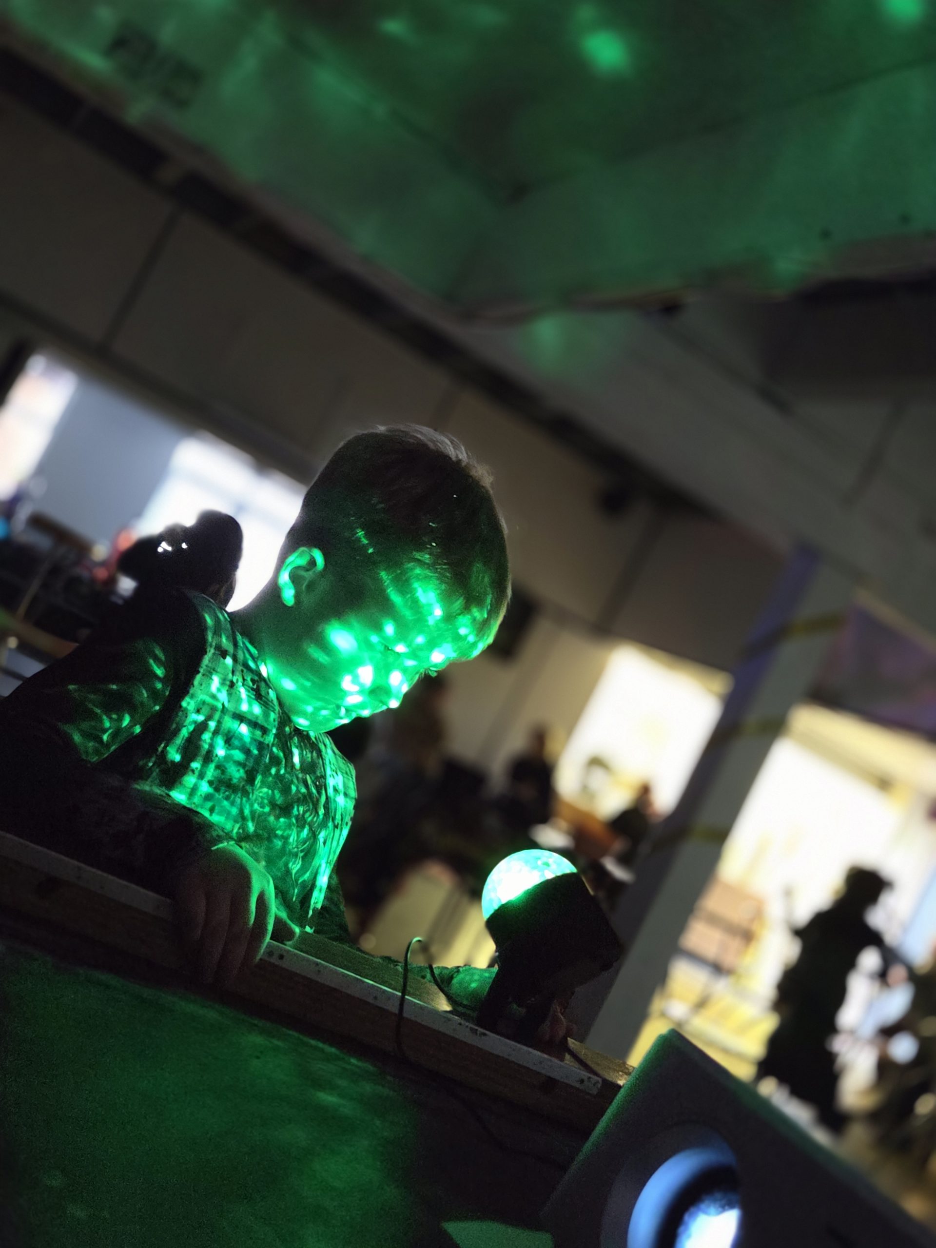 A child explores sensory lighting in a darkened room, with green light patterns projected across their face and clothing. A small glowing light sits nearby, creating a calm, immersive sensory environment.