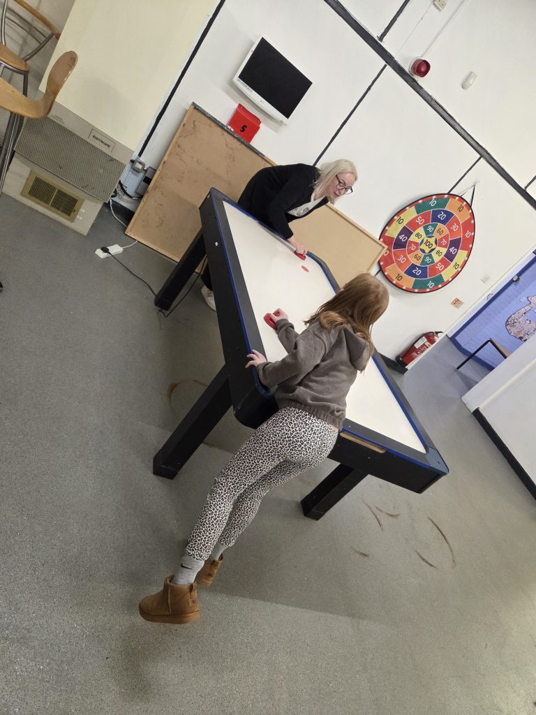 **Alternative text:** A child and an adult playing air hockey together in an indoor activity room. A large target board, fire extinguisher, chairs and wall-mounted equipment are visible in the background.