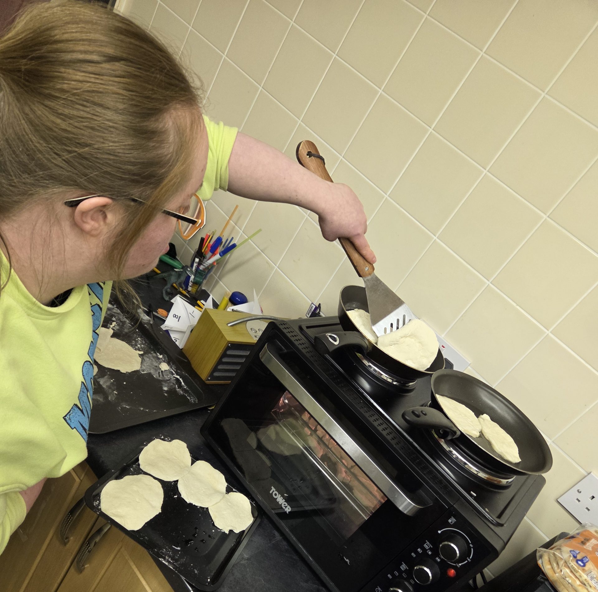 An adult cooking in a kitchen, helping pour mixture into a pan on a hot plate. Pancakes or flatbreads are being prepared, with utensils and ingredients nearby.