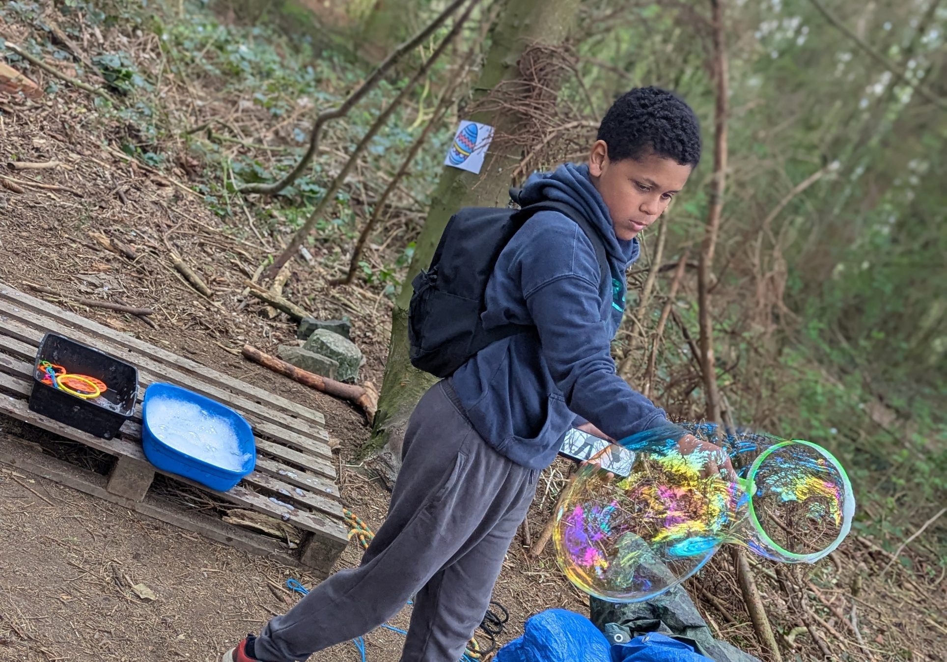 A child takes part in an outdoor nature activity beside trees and leaves, using a net and equipment to explore the area. A container and activity materials are placed nearby on the ground.