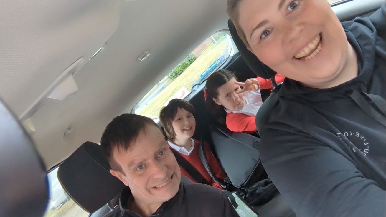 A group selfie taken inside a car, showing two adults in the front and two children in the back seats. Everyone is smiling or posing, and the children appear to be wearing red and white school uniforms.