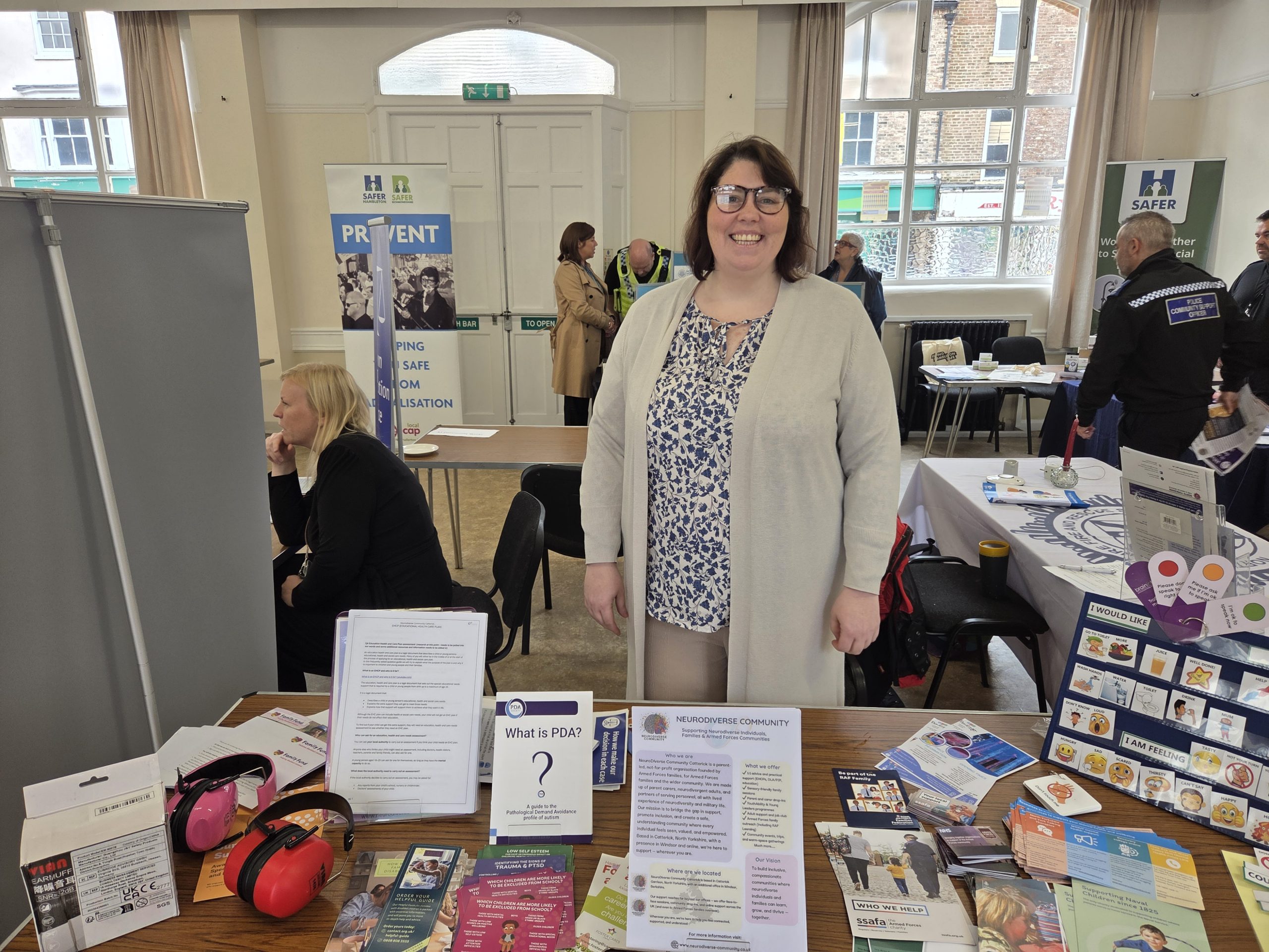 **Alternative text:** A person smiling behind a NeuroDiverse Community information stall at a community safety event. The table displays leaflets, visual communication resources, sensory ear defenders, PDA information and support materials for neurodivergent individuals, families and Armed Forces communities. Other organisation stands and attendees are visible in the background.