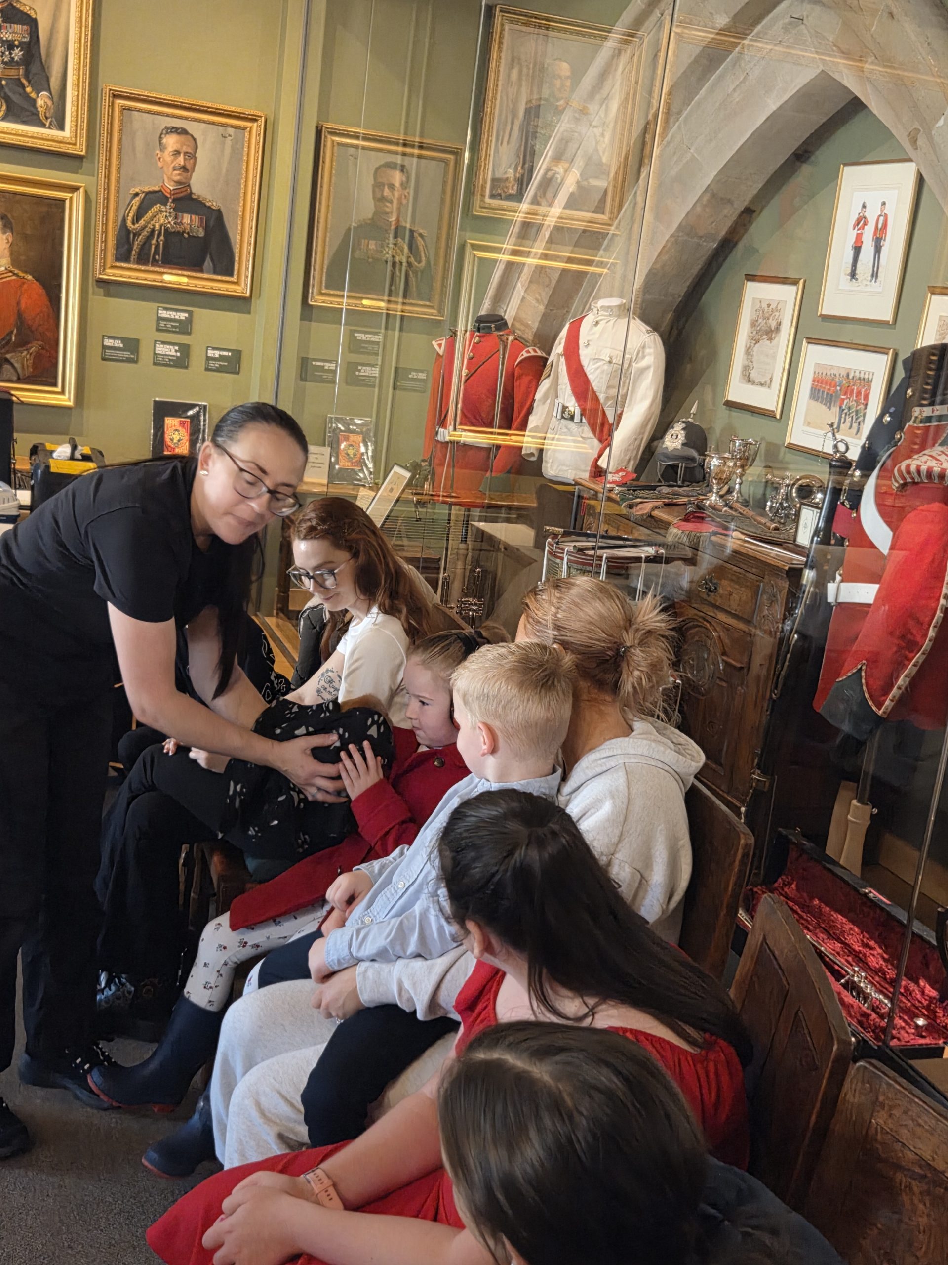 **Alternative text:** A group of children and adults sitting together in a military museum gallery, looking at and touching a soft item being shown by a staff member. Historic uniforms, medals, portraits and display cabinets are visible in the background.