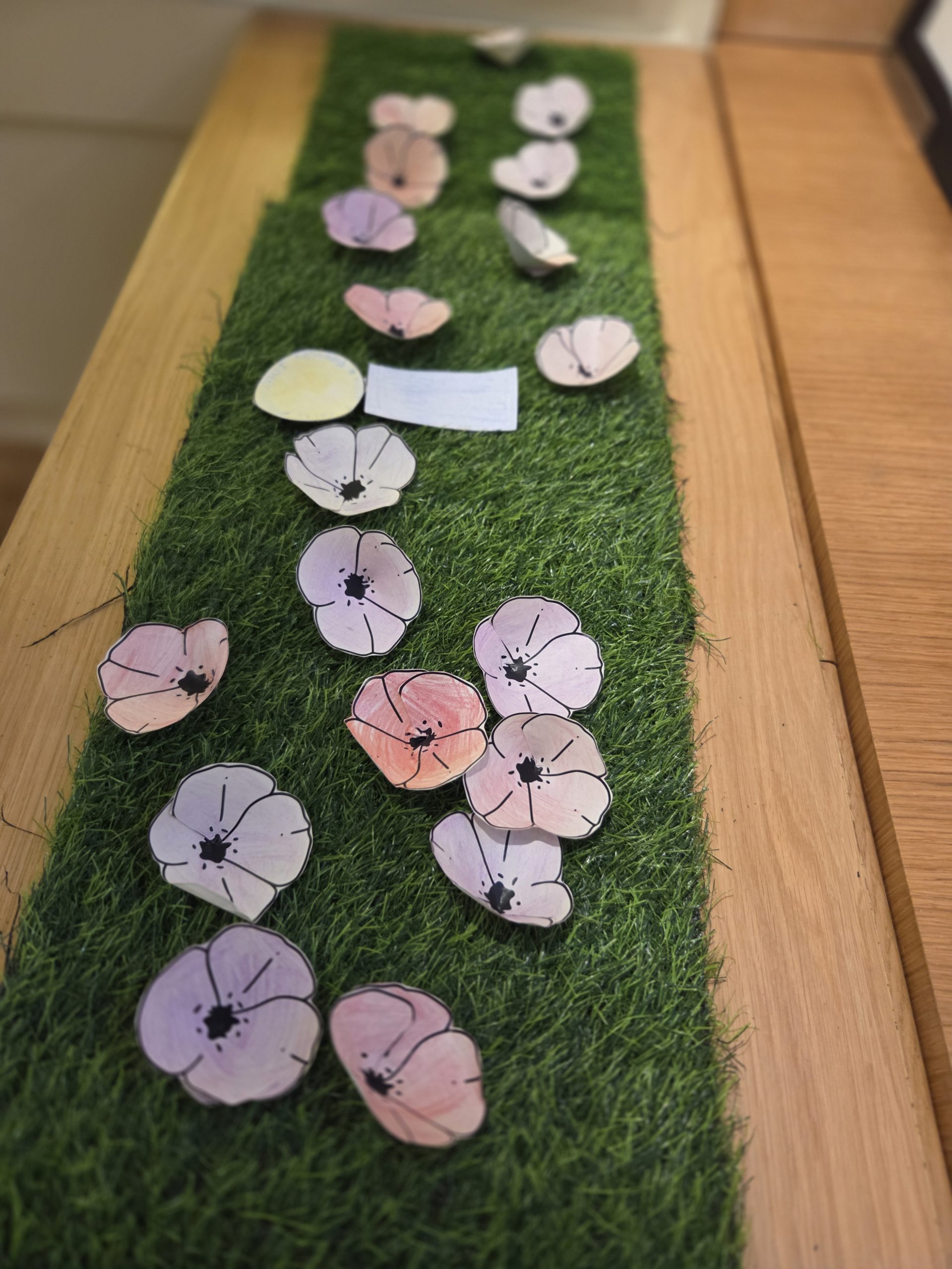 **Alternative text:** Hand-coloured paper poppies arranged on a strip of artificial grass on a wooden surface, creating a Remembrance-themed craft display.