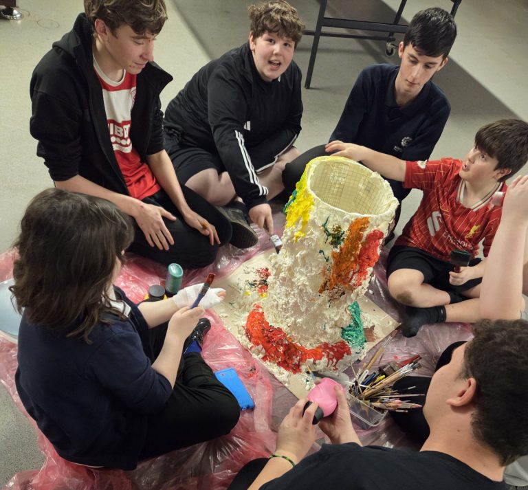 **Alternative text:** A group of children and young people sitting on the floor around a large papier-mâché volcano model, painting it with bright colours. Paintbrushes, paints and a protective sheet are spread out around them during the group craft activity.