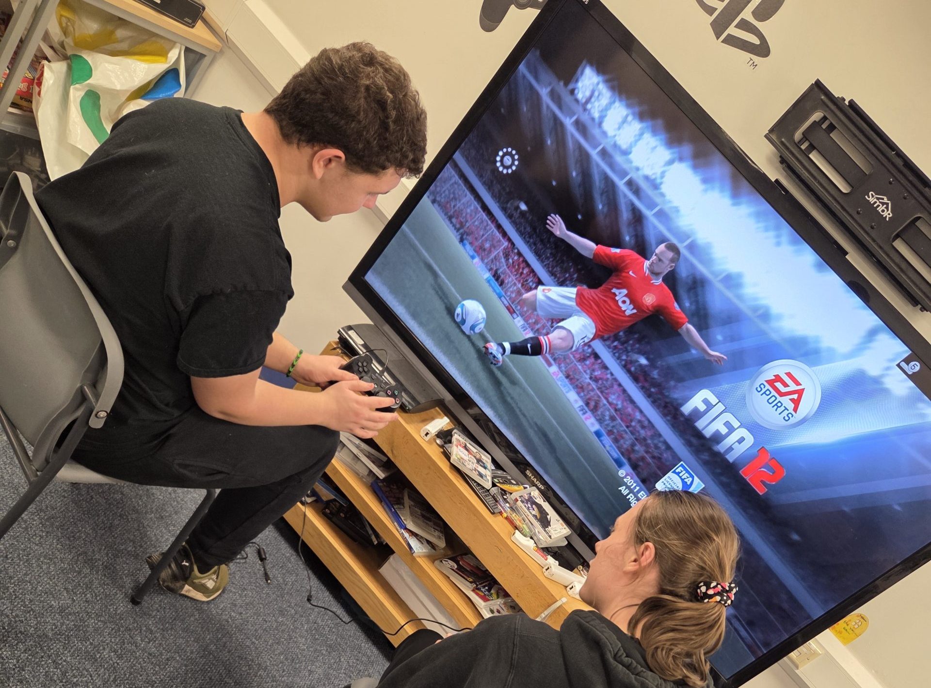 **Alternative text:** Two young people sitting indoors playing a football video game on a large TV screen using game controllers. The room includes gaming equipment, shelves, a PlayStation logo on the wall and other activity resources.