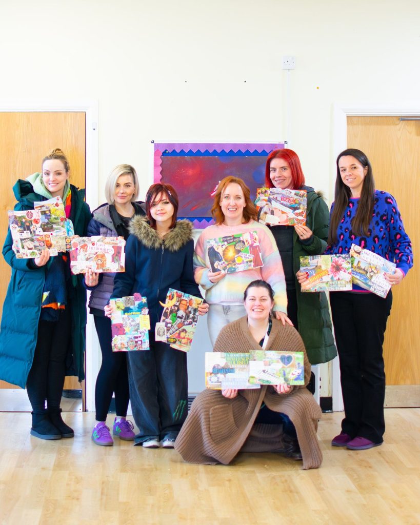 A group of seven people posing indoors in a community room, smiling and holding colourful collage artwork or vision boards. The room has wooden doors, a light wooden floor, and a noticeboard in the background.