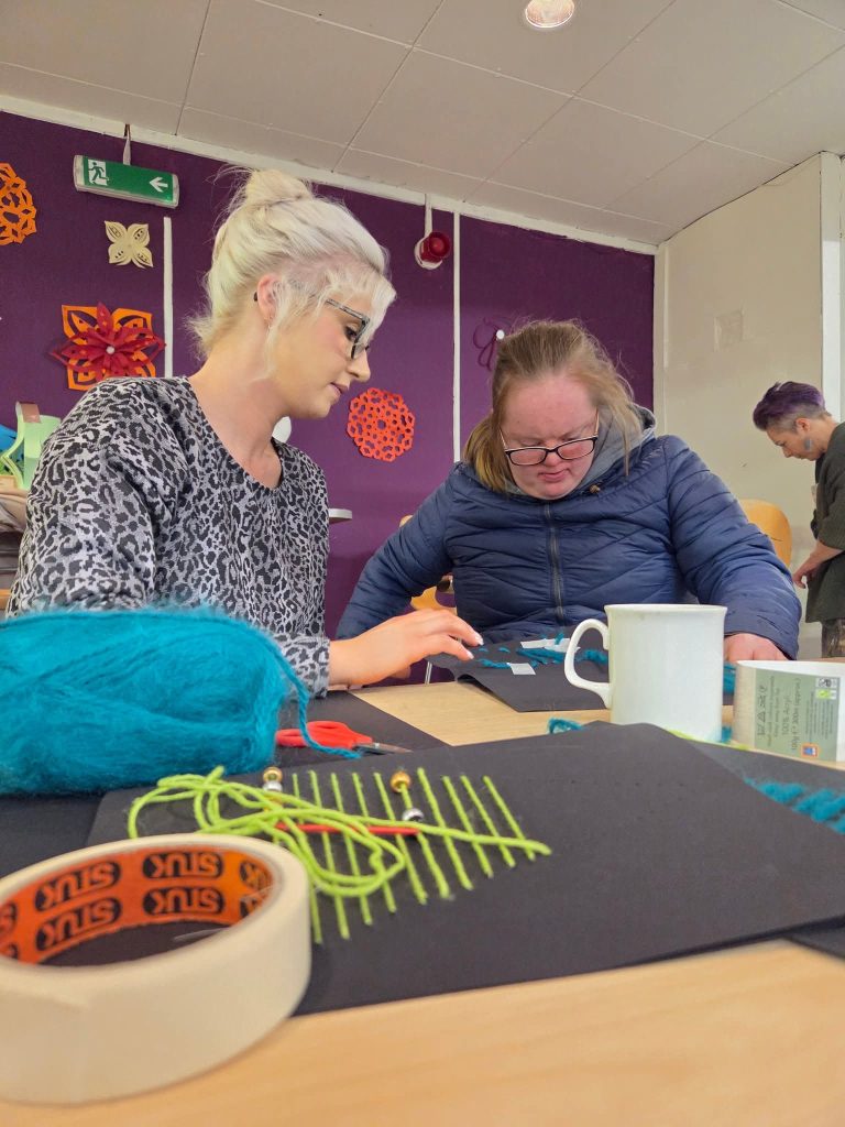 **Alternative text:** People taking part in a craft activity around a table, using wool, card, tape and scissors. One person is supporting another with a threading or weaving task, while another person works in the background. The room has purple walls with colourful decorations.