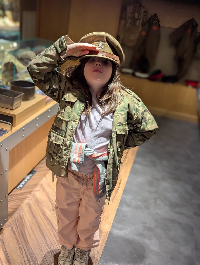 A child standing indoors in a museum or military-themed display area, wearing an oversized camouflage jacket and a military-style cap while saluting. The child is also wearing a white top, pink trousers, trainers, and a jumper tied around their waist. Display cabinets and military items are visible in the background.