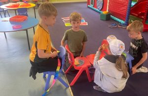 Children sitting together on small chairs and on the floor in an indoor play area, eating snacks and socialising. Colourful tables, craft materials and a soft play structure with a red slide are visible in the background.
