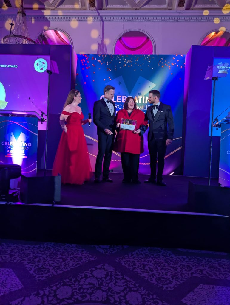 **Alternative text:** A person standing on stage at the Celebrating Forces Families Awards, holding an award certificate while three presenters stand beside them. The stage is lit with purple and blue lighting, with event branding, screens and formal decorations in the background.