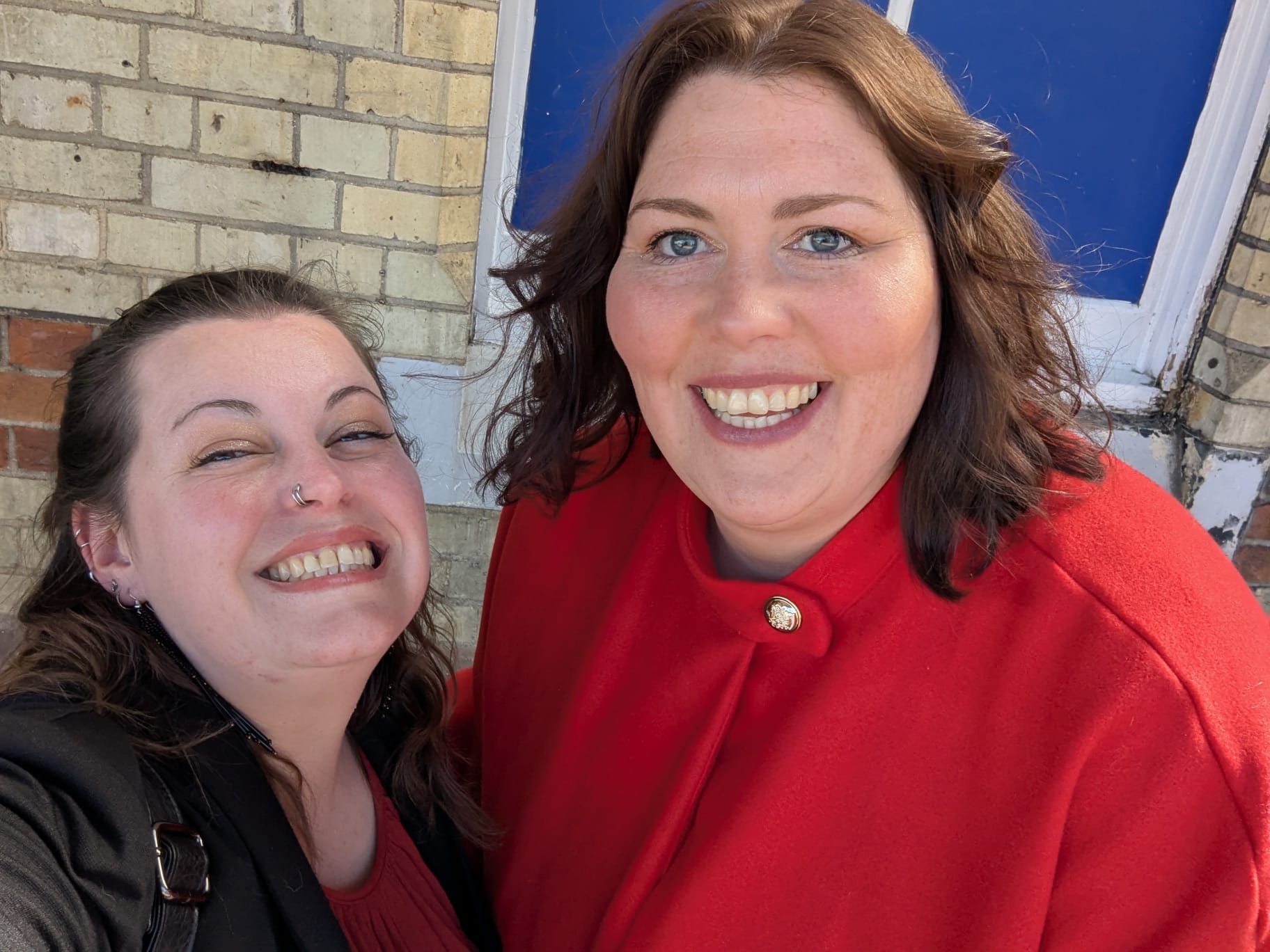 **Alternative text:** Two women smiling together for a selfie outside a brick building with a blue door. One is wearing a black jacket and red top, and the other is wearing a bright red coat.
