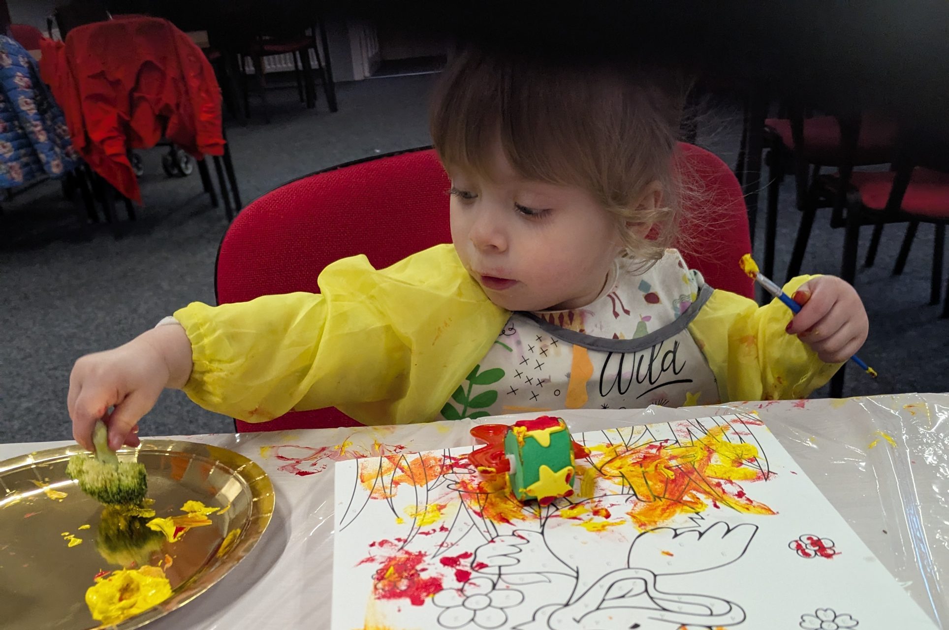 A young child wearing a yellow painting apron sits at a table doing a messy painting activity. They are using broccoli and a paintbrush to apply yellow, orange and red paint onto a colouring sheet, with paint trays and protective table covering visible around them.