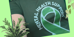 A smiling person taking a selfie, wearing a dark green “Mental Health Support” T-shirt with a green ribbon design. The image has a light green background with decorative leafy illustrations around the edges.