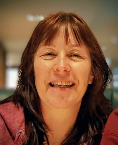 A woman with dark hair and a fringe smiles at the camera in a close-up portrait photo, with a softly blurred indoor background behind her.