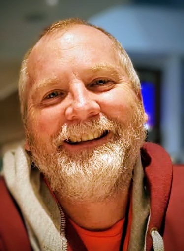 A smiling man with a beard wearing a red hoodie looks at the camera in a close-up portrait photo, with a softly blurred indoor background behind him.