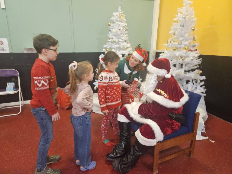 Children stand indoors in a festive Christmas setting, taking turns to speak with Santa, who is seated beside two white decorated Christmas trees. An adult dressed as an elf supports the children during the visit, with wrapped gifts and Christmas decorations nearby.
