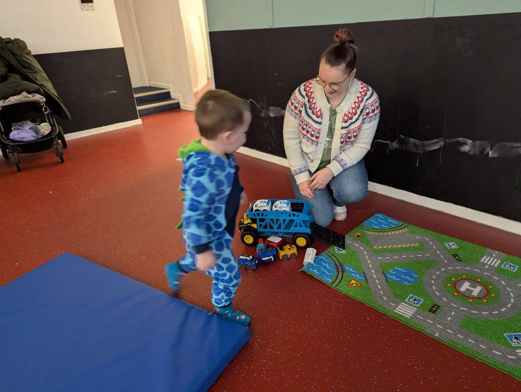 A young child plays indoors with toy vehicles beside a road-pattern play mat, while an adult kneels nearby and watches. A blue soft mat is on the floor, creating a relaxed early years play area.