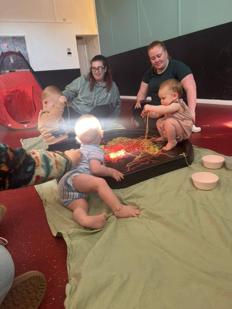 Babies and adults sit together on the floor during a sensory play session, exploring colourful spaghetti in a large tray with lights shining through it. The group is gathered on soft blankets in an indoor community space, with bowls and a small play tent nearby.