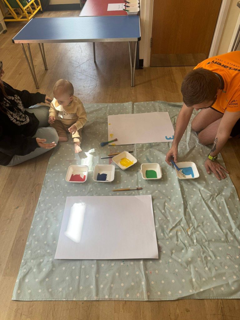A baby and an adult sit on a protective floor mat during a painting activity. Pots of colourful paint, brushes and large sheets of white paper are laid out on the mat, while the adult helps prepare blue paint for the activity.