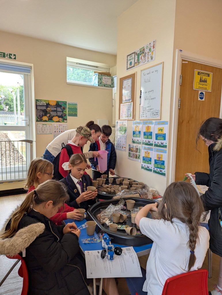 Children and volunteers take part in an indoor Eco Forces activity, filling small pots with soil and preparing seeds at tables inside a community room. Posters, a whiteboard and gardening displays are visible on the walls, creating a hands-on learning environment focused on nature, planting and exploration.