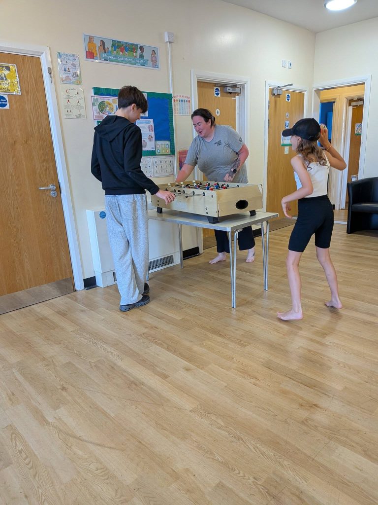 Two children and an adult play table football indoors in a bright community room, while another child stands nearby holding a ball and watching the game. The room has wooden flooring, noticeboards and classroom-style doors in the background.