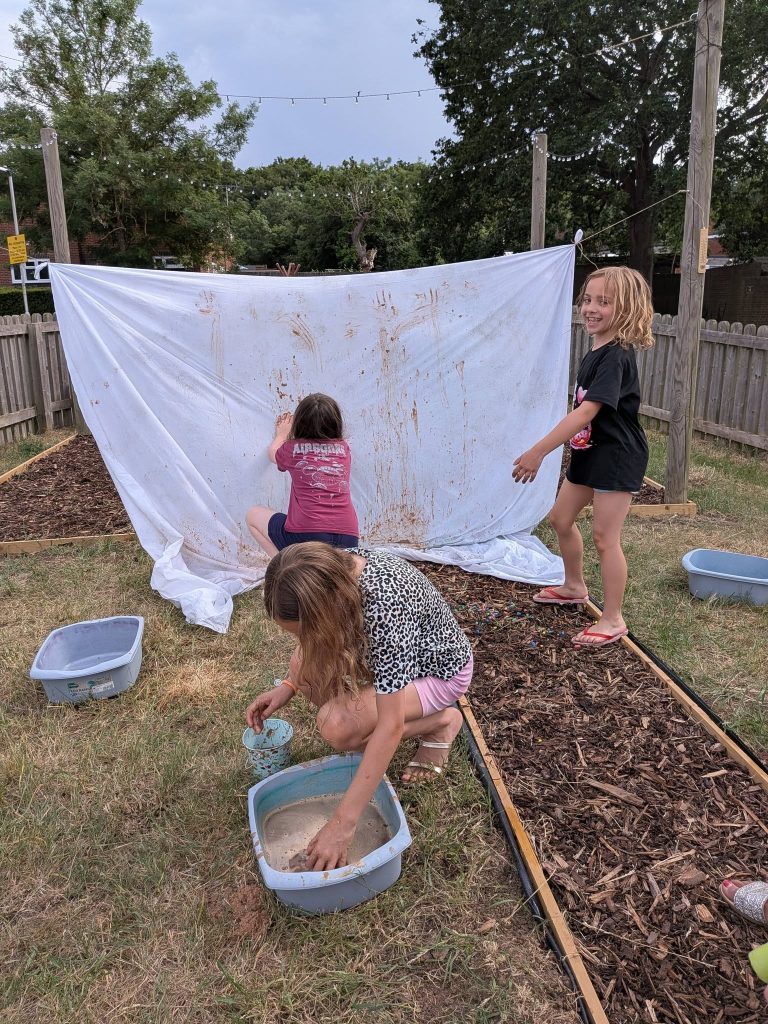 Three children take part in an outdoor messy play activity in a garden area, using muddy water to make handprints and marks on a large white sheet hanging between posts. Bowls of water and mud are on the grass, and one child smiles towards the camera while the others continue playing.