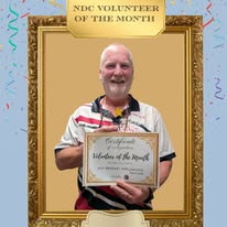 A Volunteer of the Month graphic for NDC, showing a smiling man standing inside a decorative gold frame and holding a certificate. The background is light blue with small colourful celebration details.