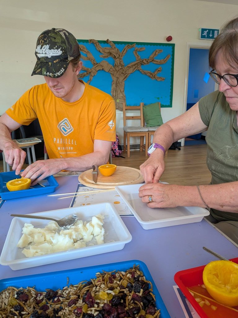 Two adults sit at a table during a hands-on activity, preparing orange halves and bird feeder ingredients. Trays contain seeds, dried fruit, mealworms and soft food mixture, with skewers and instructions on the table. A craft tree display is visible on the wall behind them.