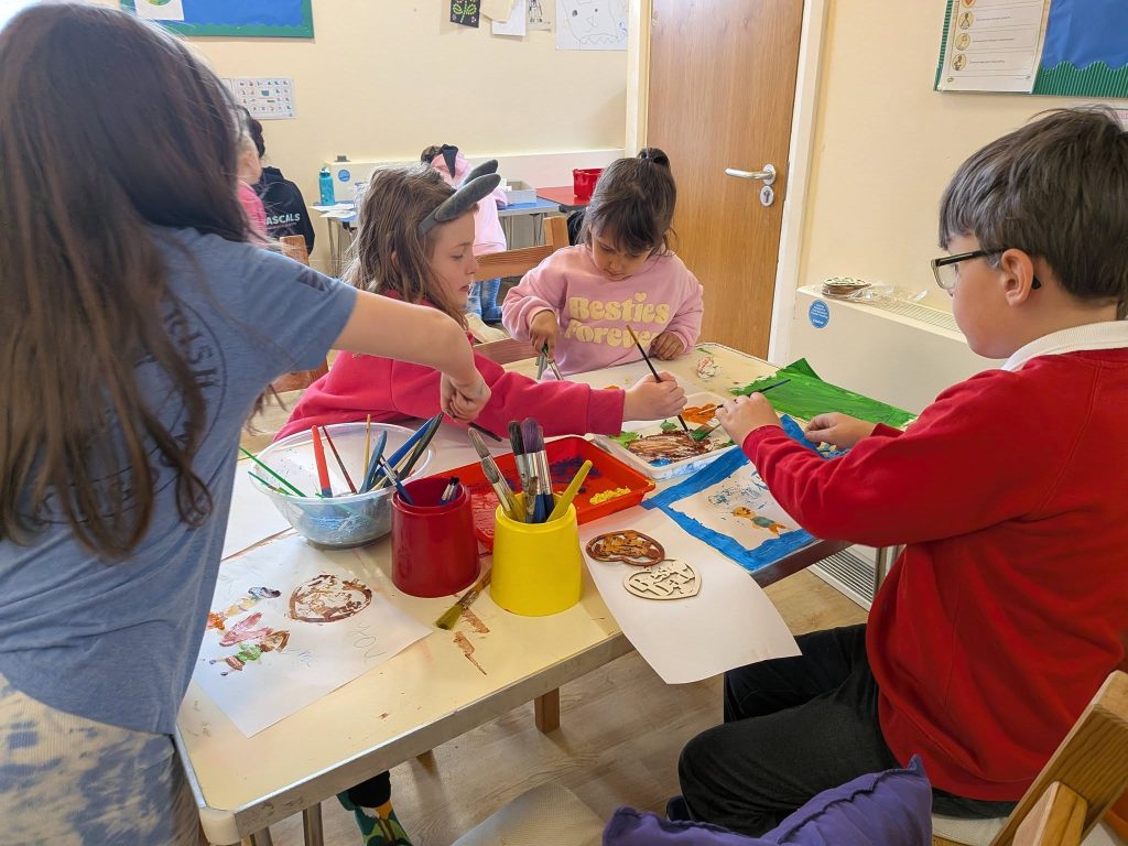 Children sit around a table indoors, painting and creating artwork together with brushes, paper and colourful paints. Pots of brushes and trays of paint are spread across the table during the creative group activity.