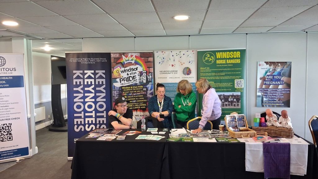 People stand behind an information stall at an indoor community event, with leaflets, resources and display materials laid out across the tables. Banners behind the stall include NeuroDiverse Community CIC, Windsor & Eton Pride, Windsor Horse Rangers and other local organisations.
