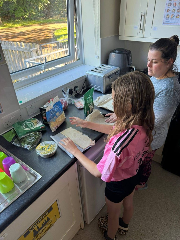 An adult and a child stand at a kitchen counter preparing food together. Pastry sheets and ingredients are laid out on the worktop, including cheese, spinach, pasta and other packets, with a toaster and air fryer beside the window.
