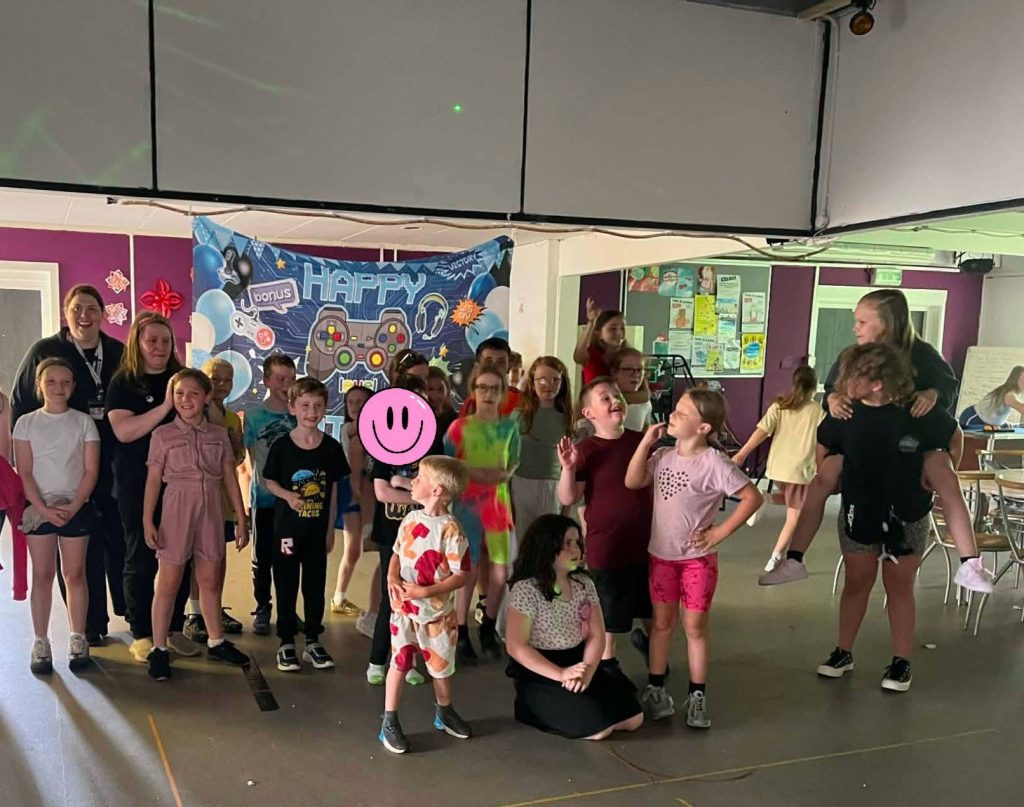 A large group of children, young people and adults gather indoors for a group photo in front of a blue “Happy Birthday” gaming-themed backdrop. Some children are smiling, posing, chatting or being held, while one person’s face is covered with a pink smiley sticker for privacy. The room has purple walls, chairs and tables to the side, and a bright, lively party atmosphere.