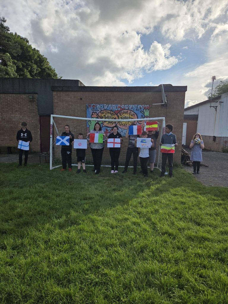 A group of children and adults stand outside on grass in front of a football goal, holding handmade country flags. Behind them is a colourful “Group Agreement” poster with messages such as “no litter, no bullying,” “have fun,” “join in,” “smile,” and “stay safe.” The scene is outdoors beside a brick building under a bright, cloudy sky.
