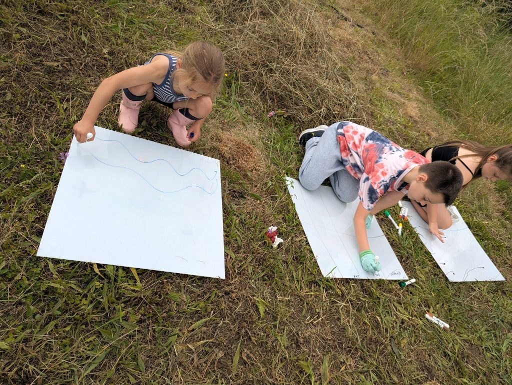Three children sit on grass outdoors, drawing and writing on large sheets of white paper with coloured markers. Pens are scattered around them, and the activity appears to be a creative outdoor group session.