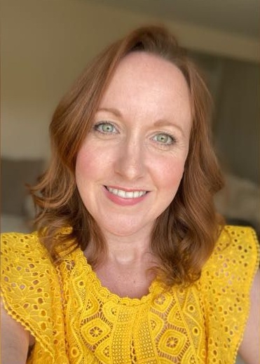 A smiling woman with red hair wearing a bright yellow lace top looks at the camera in a close-up portrait photo, with a softly blurred indoor background behind her.