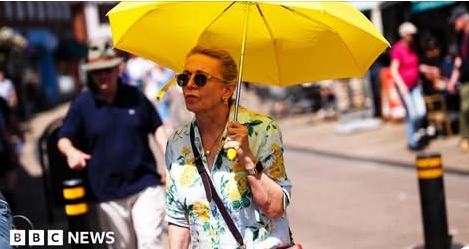 A person wearing sunglasses walks outdoors in bright sunshine while holding a yellow umbrella for shade. People are visible in the background, and a BBC News logo appears in the corner of the image.