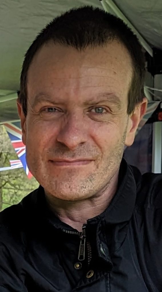 A man wearing a dark jacket smiles slightly in a close-up portrait photo taken outdoors under a canopy, with bunting visible in the background.