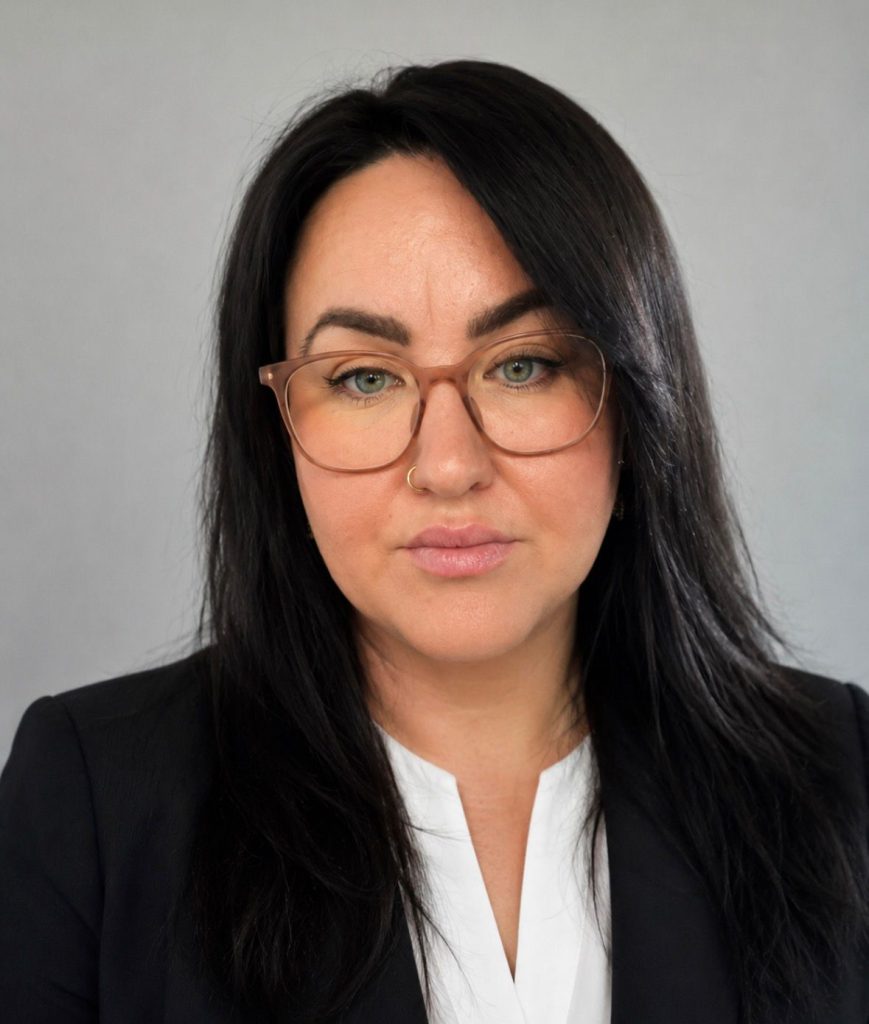 Professional headshot of a woman with long dark hair wearing glasses, a black blazer and a white blouse, looking directly at the camera against a plain grey background.