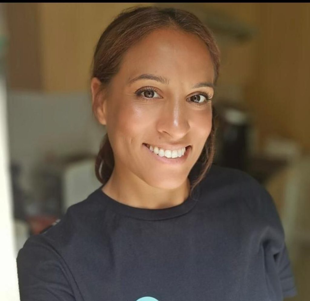 A smiling woman with brown hair tied back, wearing a dark top, looks at the camera in a close-up portrait photo with a softly blurred indoor background behind her.