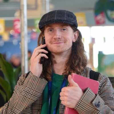 A smiling man with long hair wearing a flat cap and checked jacket holds a phone to his ear while giving a thumbs up and carrying a red folder in a busy indoor setting.
