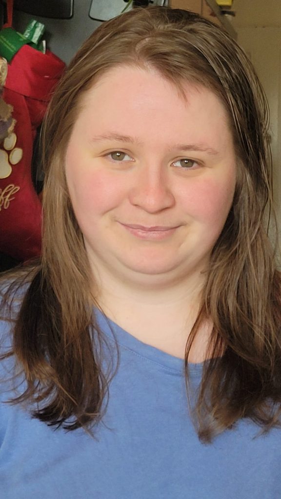 A young woman with long brown hair wearing a blue top looks directly at the camera in a close-up portrait photo, smiling softly indoors.
