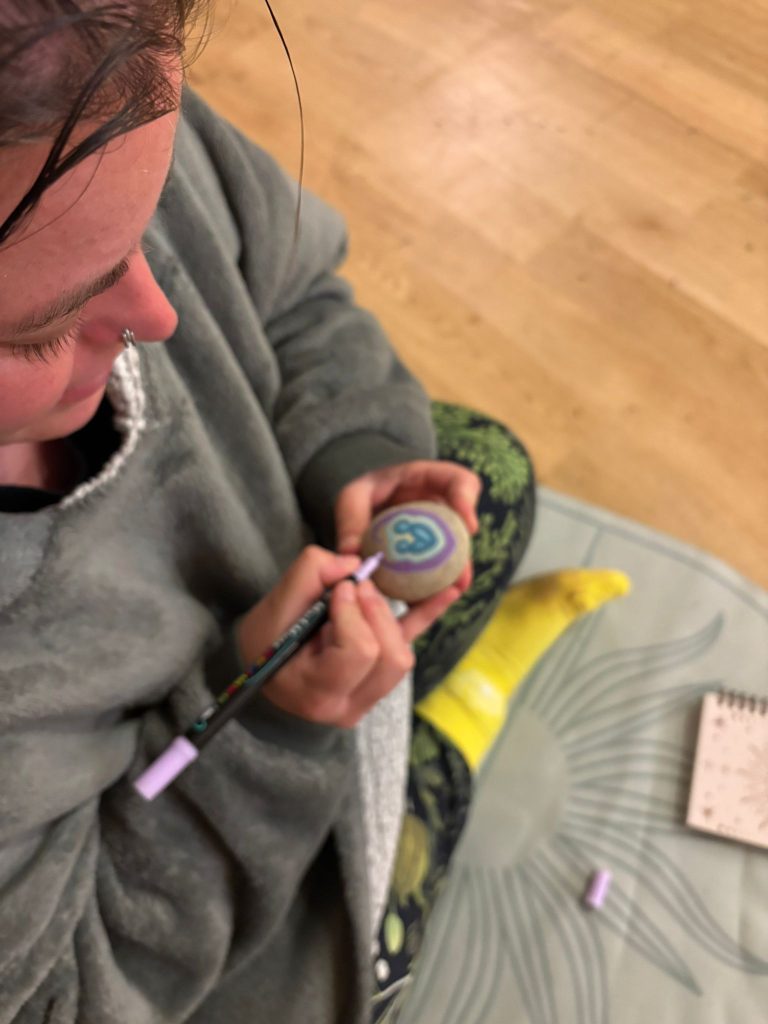 A person sits on the floor during a calming creative activity, drawing a purple and blue design onto a smooth stone with a marker pen. A notebook and pen lid are nearby on a soft mat.