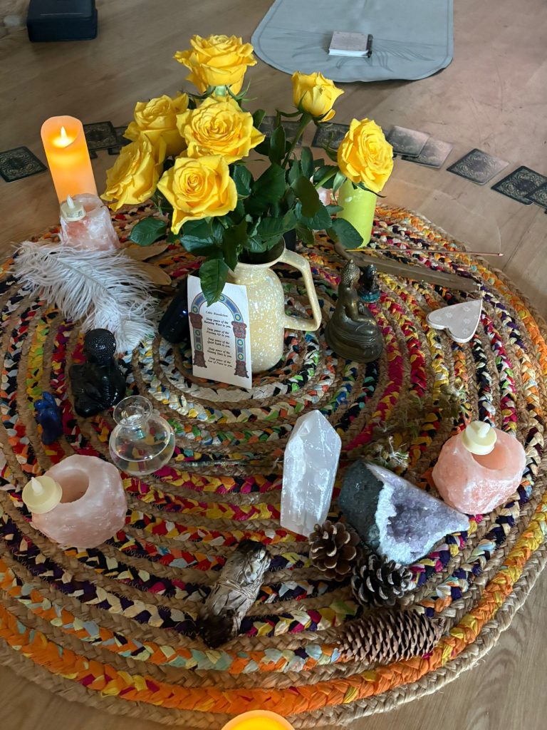 A calming wellbeing centrepiece arranged on a colourful woven circular rug, featuring yellow roses in a jug, LED candles, crystals, pinecones, feathers, small ornaments and cards placed around the edge.