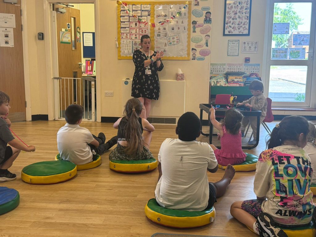 Children sit on floor cushions in a community room while an adult leads a BSL learning activity at the front. The children are watching, joining in with signs and taking part in an inclusive group session with visual learning resources and posters displayed around the room.