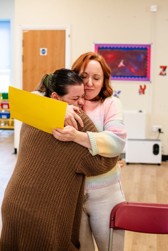 Two adults share an emotional hug inside a community room, with one person holding a yellow piece of paper. The image captures a supportive and caring moment, reflecting connection, reassurance and community support.