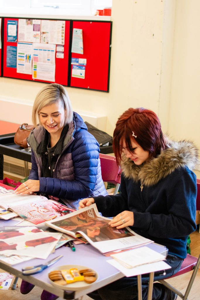 Two people sit at a table in a community room, smiling while looking through magazines and printed materials during a creative activity. Papers, scissors, pens and biscuits are spread across the table, creating a relaxed and friendly group setting.