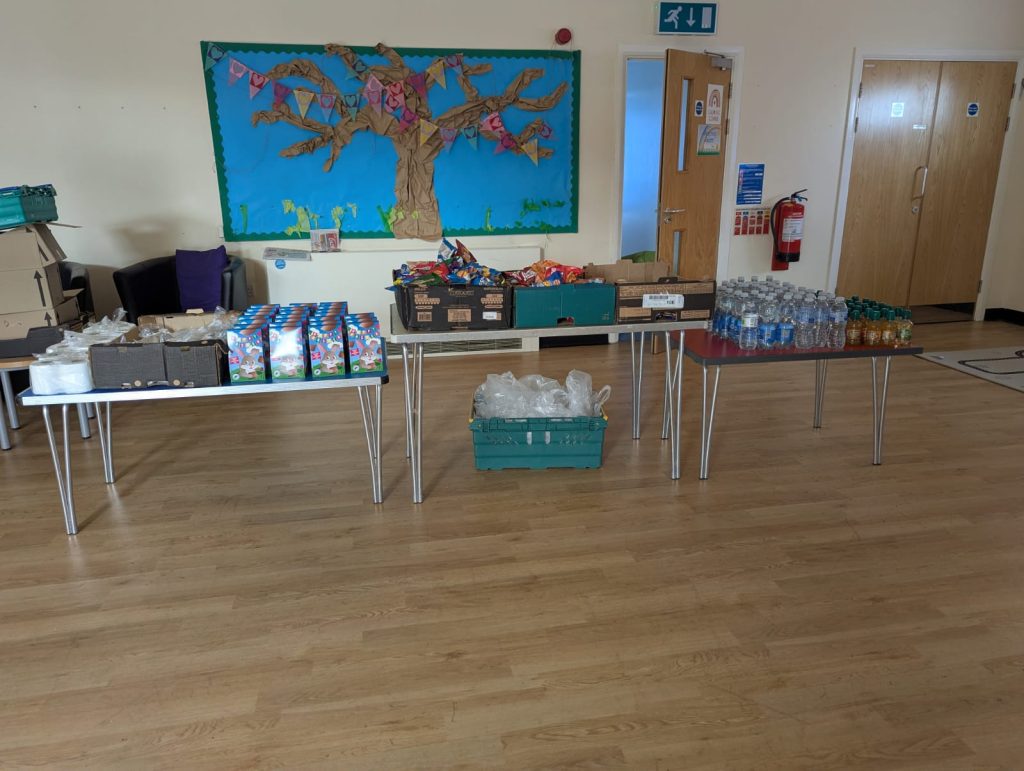 Tables in a community hall are set up with donated food, drinks and essential items, including bottled water, juice, snacks and other supplies, ready for families to access as part of a free poverty relief support session.